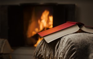 Peceful image of open book resting on a arm rest of a couch. Warm fireplace on background.